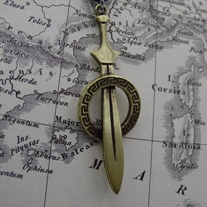 Może przedstawiać: Złoty wisiorek w kształcie miecza z okrągłym, ozdobnym wzorem w stylu greckiej klucza. Wisiorek znajduje się na mapie starożytnej Europy.