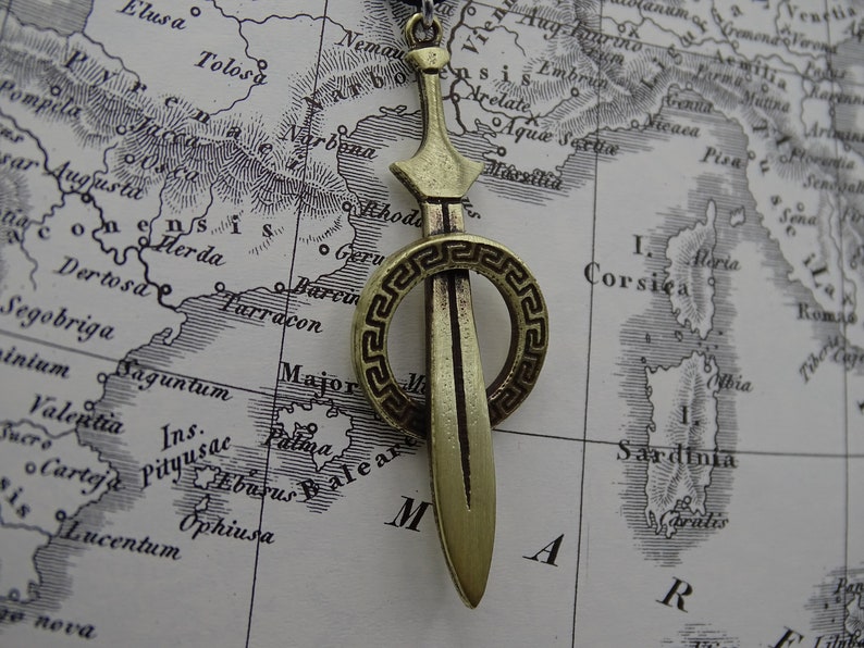 Może przedstawiać: Złoty wisiorek w kształcie miecza z okrągłym, ozdobnym wzorem w stylu greckiej klucza. Wisiorek znajduje się na mapie starożytnej Europy.