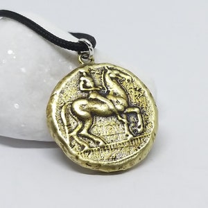 Puede incluir: Un collar con un colgante redondo en forma de moneda dorada que representa un caballo y su jinete en relieve. El colgante est&aacute; en un cord&oacute;n negro.