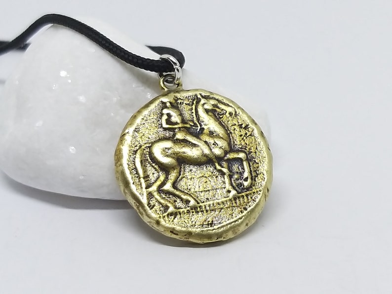 Puede incluir: Un collar con un colgante redondo en forma de moneda dorada que representa un caballo y su jinete en relieve. El colgante est&aacute; en un cord&oacute;n negro.