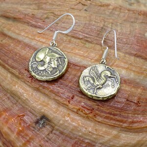 May include: A pair of gold-toned earrings with a silver hook. One earring features a winged horse, the other a profile of a woman wearing a helmet.