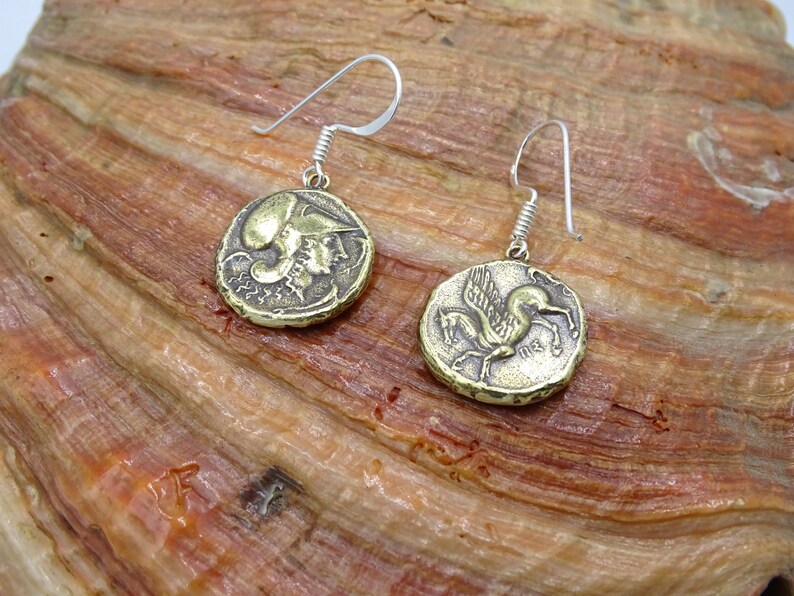 May include: A pair of gold-toned earrings with a silver hook. One earring features a winged horse, the other a profile of a woman wearing a helmet.