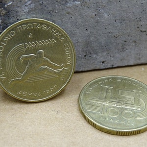 May include: Two gold-colored Greek drachma coins. The first coin features a runner with the text "XI PANATHINAIKO PROTAGMA ATHL". The second coin features a building with the text "BANK OF GREECE 100 DRACHMAE".