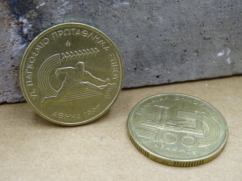 May include: Two gold-colored Greek drachma coins. The first coin features a runner with the text "XI PANATHINAIKO PROTAGMA ATHL". The second coin features a building with the text "BANK OF GREECE 100 DRACHMAE".