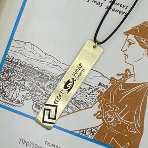 May include: A gold-coloured metal bookmark with an inscription in Greek. The bookmark features a stylised ship design and the text "&Epsilon;&Sigma;&Sigma;&Epsilon;&Tau;&Iota; &Eta;&Mu;&Alpha;&Rho; &Omicron;&Iota;&Nu;&Kappa;&Rho;&Omicron;&Sigma;". The bookmark is hanging from a black cord.