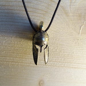 May include: A gold-colored metal pendant necklace featuring a detailed Spartan helmet design. The necklace is on a black cord.
