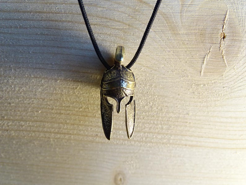 May include: A gold-colored metal pendant necklace featuring a detailed Spartan helmet design. The necklace is on a black cord.