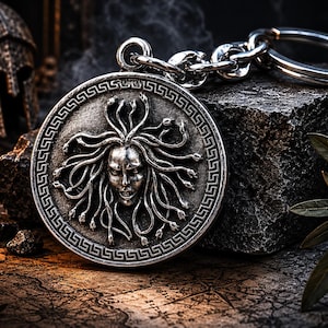 Op de afbeelding: Een zilverkleurige sleutelhanger met een gedetailleerd Medusa-hoofdontwerp. De ronde hanger heeft een Griekse sleutelrand en een ketting met een sleutelring. De achtergrond omvat een stenen oppervlak en een kaart.
