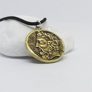 May include: A gold-colored necklace with a round pendant featuring a detailed profile of a man with a laurel wreath and the inscription "PANEION".
