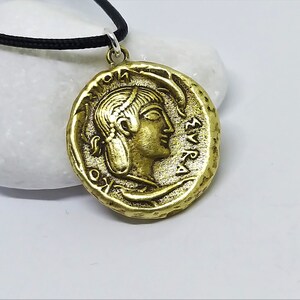 Puede incluir: Un collar con un colgante en forma de moneda griega antigua de color dorado, que representa el perfil de una mujer y letras griegas. El colgante est&aacute; en un cord&oacute;n negro.