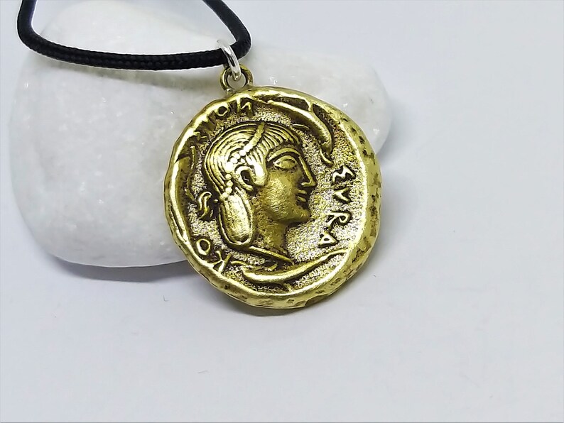 Puede incluir: Un collar con un colgante en forma de moneda griega antigua de color dorado, que representa el perfil de una mujer y letras griegas. El colgante est&aacute; en un cord&oacute;n negro.