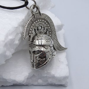 Könnte beinhalten: Silberner Anhänger mit einer detaillierten Darstellung eines griechischen Kriegerhelms mit einer Feder. Der Helm hat komplizierte Designs und eine strukturierte Oberfläche.
