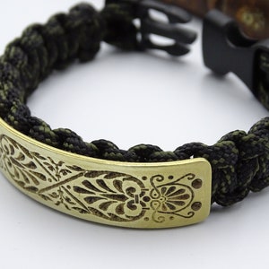 Puede incluir: Pulsera trenzada de paracord en negro y verde oliva con una placa decorativa de lat&oacute;n. La placa de lat&oacute;n presenta un dise&ntilde;o floral grabado y se asegura con un cierre de pl&aacute;stico negro.