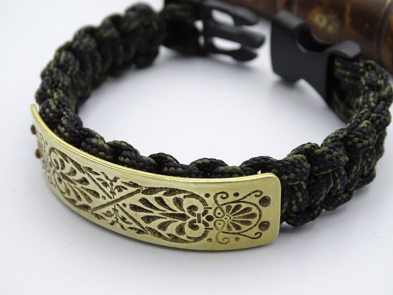 Puede incluir: Pulsera trenzada de paracord en negro y verde oliva con una placa decorativa de lat&oacute;n. La placa de lat&oacute;n presenta un dise&ntilde;o floral grabado y se asegura con un cierre de pl&aacute;stico negro.