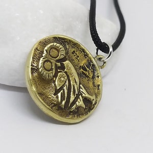 May include: A gold-colored pendant necklace featuring a round, detailed replica of an ancient Greek coin with an owl design. The necklace has a black cord.