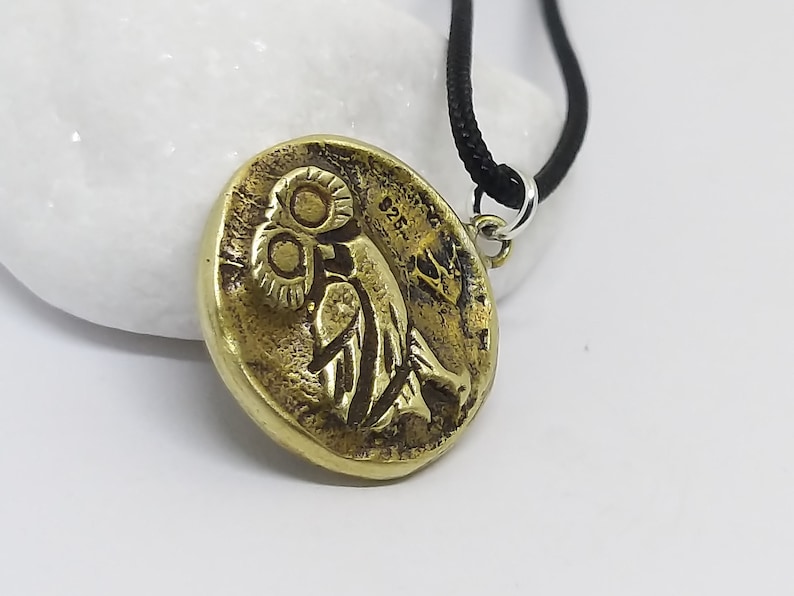 May include: A gold-colored pendant necklace featuring a round, detailed replica of an ancient Greek coin with an owl design. The necklace has a black cord.