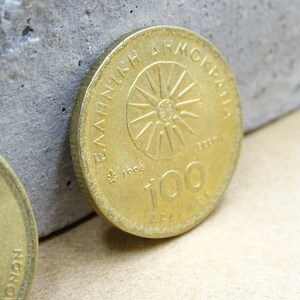 Puede incluir: Una moneda griega de color dorado con el texto "&Epsilon;&Lambda;&Lambda;&Eta;&Nu;&Iota;&Kappa;&Eta; &Delta;&Eta;&Mu;&Omicron;&Kappa;&Rho;&Alpha;&Tau;&Iota;&Alpha;" y "100 &Delta;&Rho;&Alpha;&Chi;&Mu;&Epsilon;&Sigma;" alrededor de un dise&ntilde;o de sol.