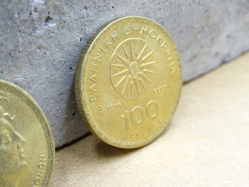 Puede incluir: Una moneda griega de color dorado con el texto "&Epsilon;&Lambda;&Lambda;&Eta;&Nu;&Iota;&Kappa;&Eta; &Delta;&Eta;&Mu;&Omicron;&Kappa;&Rho;&Alpha;&Tau;&Iota;&Alpha;" y "100 &Delta;&Rho;&Alpha;&Chi;&Mu;&Epsilon;&Sigma;" alrededor de un dise&ntilde;o de sol.