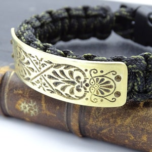 Puede incluir: Una pulsera de paracord negra y verde oliva con una placa decorativa de lat&oacute;n. La placa de lat&oacute;n presenta un dise&ntilde;o floral grabado. La pulsera tiene un cierre de pl&aacute;stico negro y est&aacute; sobre un libro antiguo.
