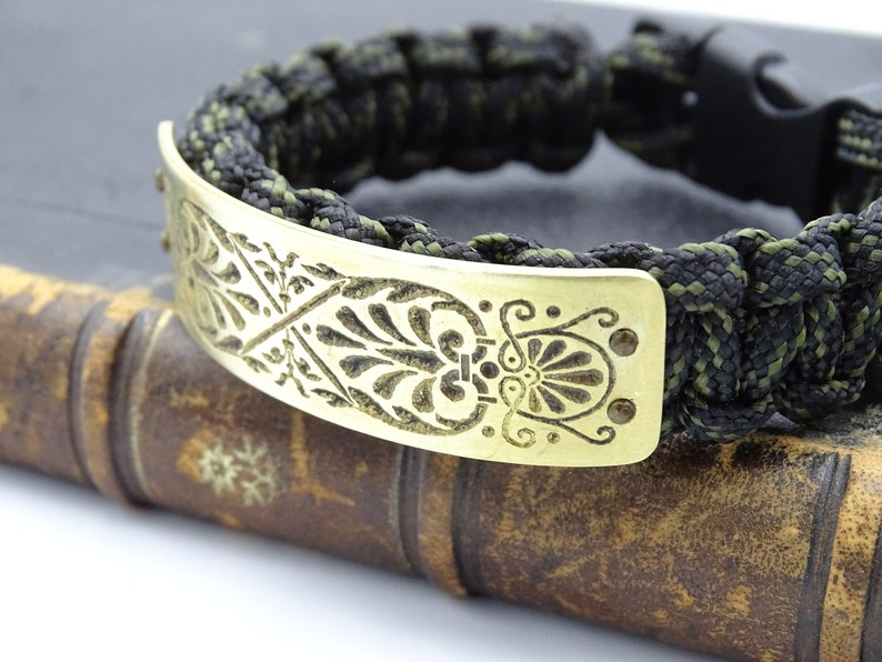 Puede incluir: Una pulsera de paracord negra y verde oliva con una placa decorativa de lat&oacute;n. La placa de lat&oacute;n presenta un dise&ntilde;o floral grabado. La pulsera tiene un cierre de pl&aacute;stico negro y est&aacute; sobre un libro antiguo.