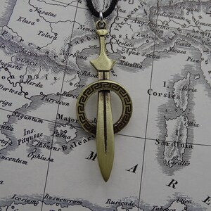 Może przedstawiać: Złoty naszyjnik z zawieszką w kształcie miecza, ozdobiony okrągłym wzorem. Miecz jest otoczony greckim wzorem klucza.