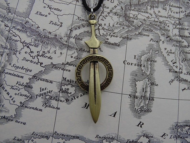 Może przedstawiać: Złoty naszyjnik z zawieszką w kształcie miecza, ozdobiony okrągłym wzorem. Miecz jest otoczony greckim wzorem klucza.