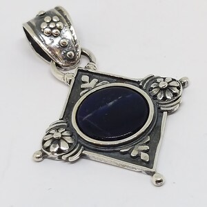 May include: A silver pendant with a dark blue gemstone set in a diamond-shaped frame with ornate floral details.