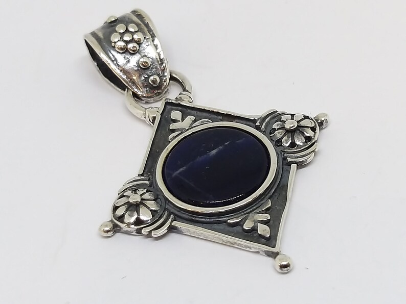 May include: A silver pendant with a dark blue gemstone set in a diamond-shaped frame with ornate floral details.