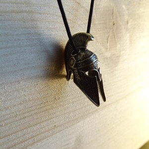 May include: A silver-toned Spartan helmet pendant on a black cord. The helmet has a detailed design with a raised crest and a pointed visor.