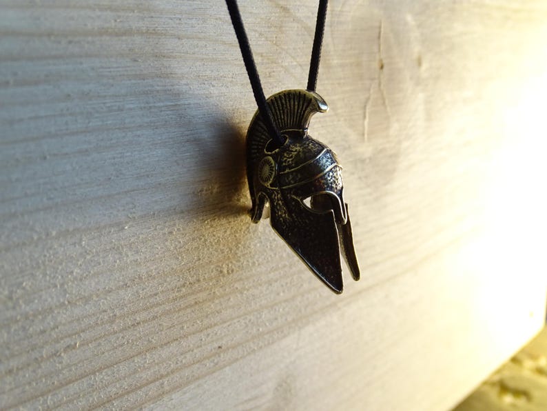 May include: A silver-toned Spartan helmet pendant on a black cord. The helmet has a detailed design with a raised crest and a pointed visor.