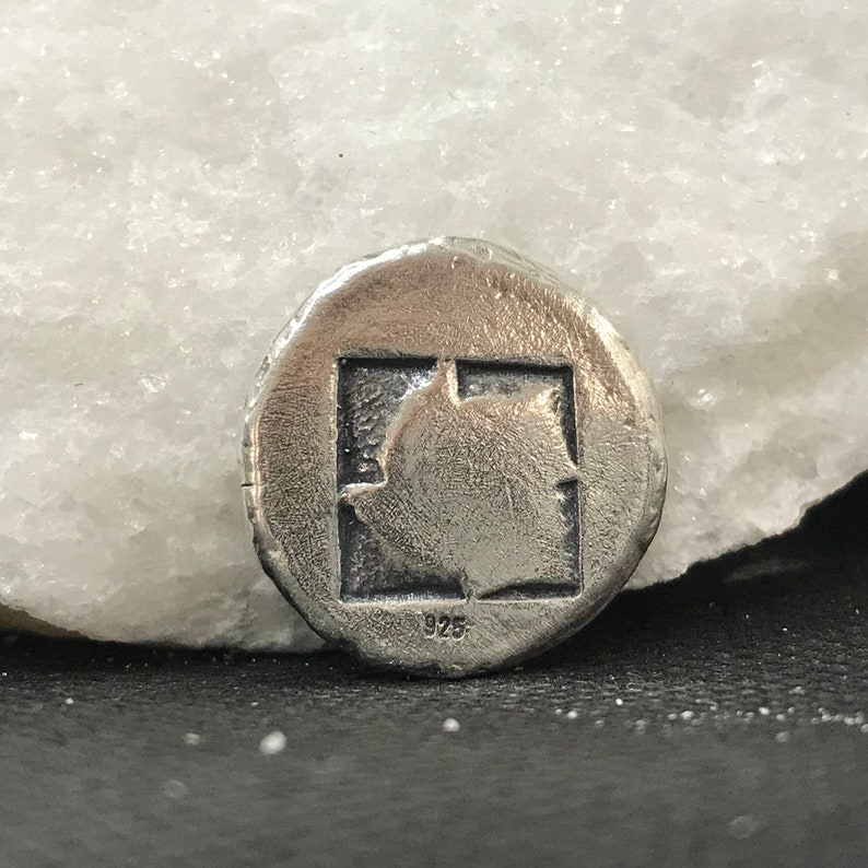 Puede incluir: Primer plano de una moneda de plata con un dise&ntilde;o cuadrado en el centro. El cuadrado contiene una imagen detallada. La moneda tiene un aspecto texturizado y envejecido. El n&uacute;mero "925" es visible en la moneda.
