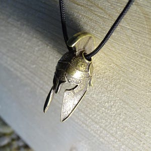 May include: A gold-toned metal pendant in the shape of a Spartan helmet. The helmet has a detailed design with a textured surface and a raised crest.