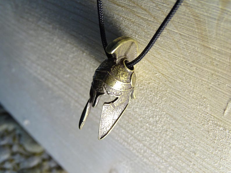 May include: A gold-toned metal pendant in the shape of a Spartan helmet. The helmet has a detailed design with a textured surface and a raised crest.