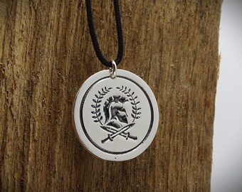 May include: Silver pendant necklace with a round medallion featuring a Spartan helmet with crossed swords and a laurel wreath. The necklace is on a black cord.