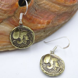 May include: A pair of gold-toned earrings featuring a round coin design with a profile of a woman wearing a helmet. The earrings are suspended from silver-toned hooks.