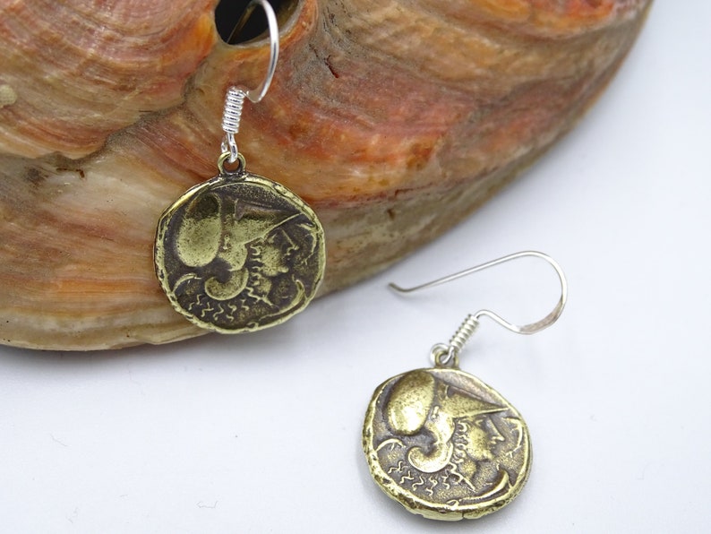 May include: A pair of gold-toned earrings featuring a round coin design with a profile of a woman wearing a helmet. The earrings are suspended from silver-toned hooks.