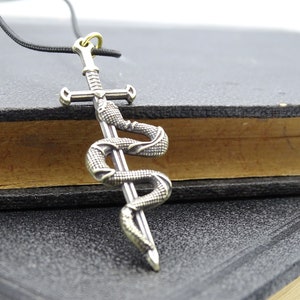 Pode incluir: Um colar com um pingente de prata que representa uma espada com uma cobra enrolada ao redor. O pingente está em um cordão preto.