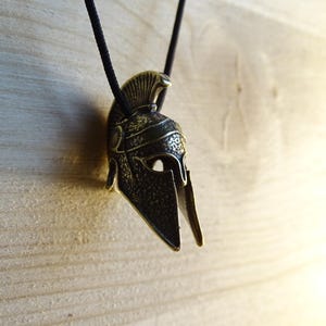 May include: A bronze-colored Spartan helmet pendant with a black cord. The helmet has a detailed design and a plume on top.