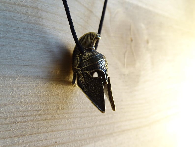 May include: A bronze-colored Spartan helmet pendant with a black cord. The helmet has a detailed design and a plume on top.