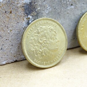 Puede incluir: Una moneda griega de color dorado con la inscripci&oacute;n "ME&Gamma;A&Sigma; &Alpha;&Lambda;&Epsilon;&Xi;&Alpha;&Nu;&Delta;&Rho;&Omicron;&Sigma; &Beta;&Alpha;&Sigma;&Iota;&Lambda;&Epsilon;&Upsilon;&Sigma;  &Mu;&Alpha;&Kappa;&Epsilon;&Delta;&Omicron;&Nu;&Omega;&Nu;" alrededor del borde y un perfil de Alejandro Magno en el centro.