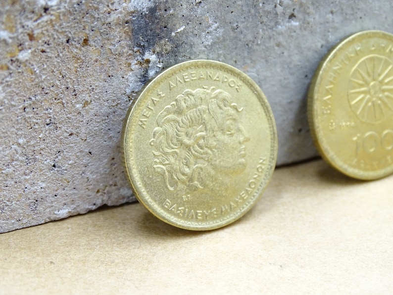 Puede incluir: Una moneda griega de color dorado con la inscripci&oacute;n "ME&Gamma;A&Sigma; &Alpha;&Lambda;&Epsilon;&Xi;&Alpha;&Nu;&Delta;&Rho;&Omicron;&Sigma; &Beta;&Alpha;&Sigma;&Iota;&Lambda;&Epsilon;&Upsilon;&Sigma;  &Mu;&Alpha;&Kappa;&Epsilon;&Delta;&Omicron;&Nu;&Omega;&Nu;" alrededor del borde y un perfil de Alejandro Magno en el centro.