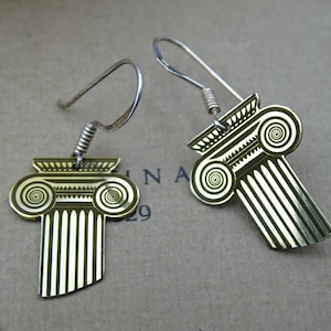 May include: A pair of gold-colored earrings shaped like Greek columns. Each earring features a detailed design with spiral accents and vertical lines, suspended from silver-toned ear wires.