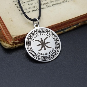 May include: A silver-colored pendant with a black cord necklace. The round pendant features Greek text and a central symbol within a decorative border. The pendant rests on a dark surface next to an open book with aged pages.