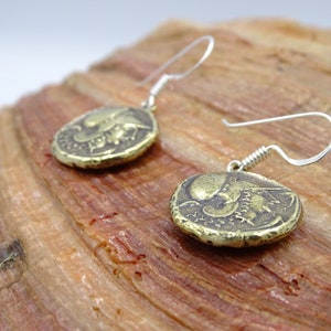 May include: A pair of gold-toned earrings with a coin-shaped design featuring a detailed image of a mythical creature. The earrings have silver-toned hooks.