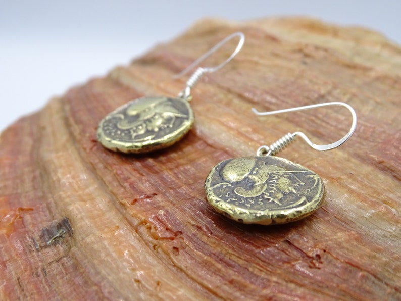 May include: A pair of gold-toned earrings with a coin-shaped design featuring a detailed image of a mythical creature. The earrings have silver-toned hooks.