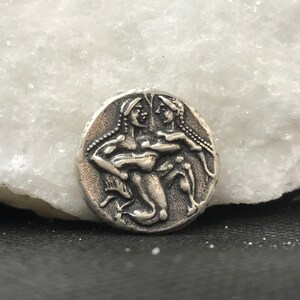 Puede incluir: Una moneda de color plateado con un relieve detallado de dos figuras, posiblemente mitol&oacute;gicas, con cabello suelto y prendas drapeadas. La moneda tiene forma circular y un aspecto ligeramente envejecido, lo que sugiere una pieza antigua o vintage.