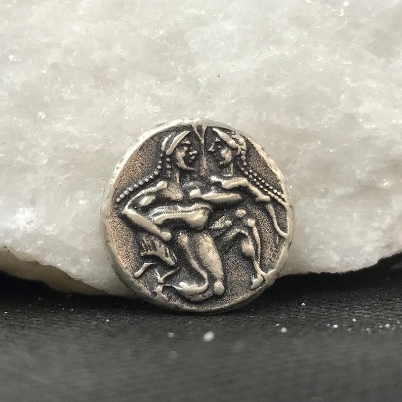 Puede incluir: Una moneda de color plateado con un relieve detallado de dos figuras, posiblemente mitol&oacute;gicas, con cabello suelto y prendas drapeadas. La moneda tiene forma circular y un aspecto ligeramente envejecido, lo que sugiere una pieza antigua o vintage.