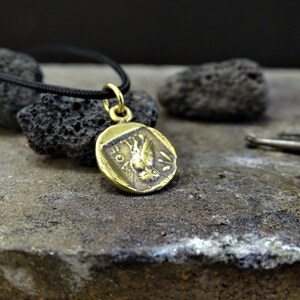 Puede incluir: Un collar de oro con un colgante redondo en forma de moneda que presenta una imagen detallada de un p&aacute;jaro con las alas extendidas. El colgante est&aacute; colgado de un cord&oacute;n negro.