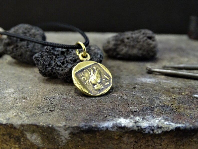 Puede incluir: Un collar de oro con un colgante redondo en forma de moneda que presenta una imagen detallada de un p&aacute;jaro con las alas extendidas. El colgante est&aacute; colgado de un cord&oacute;n negro.