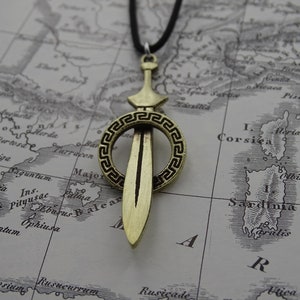 Może przedstawiać: Złoty naszyjnik z wisiorem w kształcie miecza, ozdobiony dekoracyjnym kołem. Koło ma wz&oacute;r greckiego klucza. Naszyjnik jest na czarnym sznurku i jest wyświetlany na starej mapie.